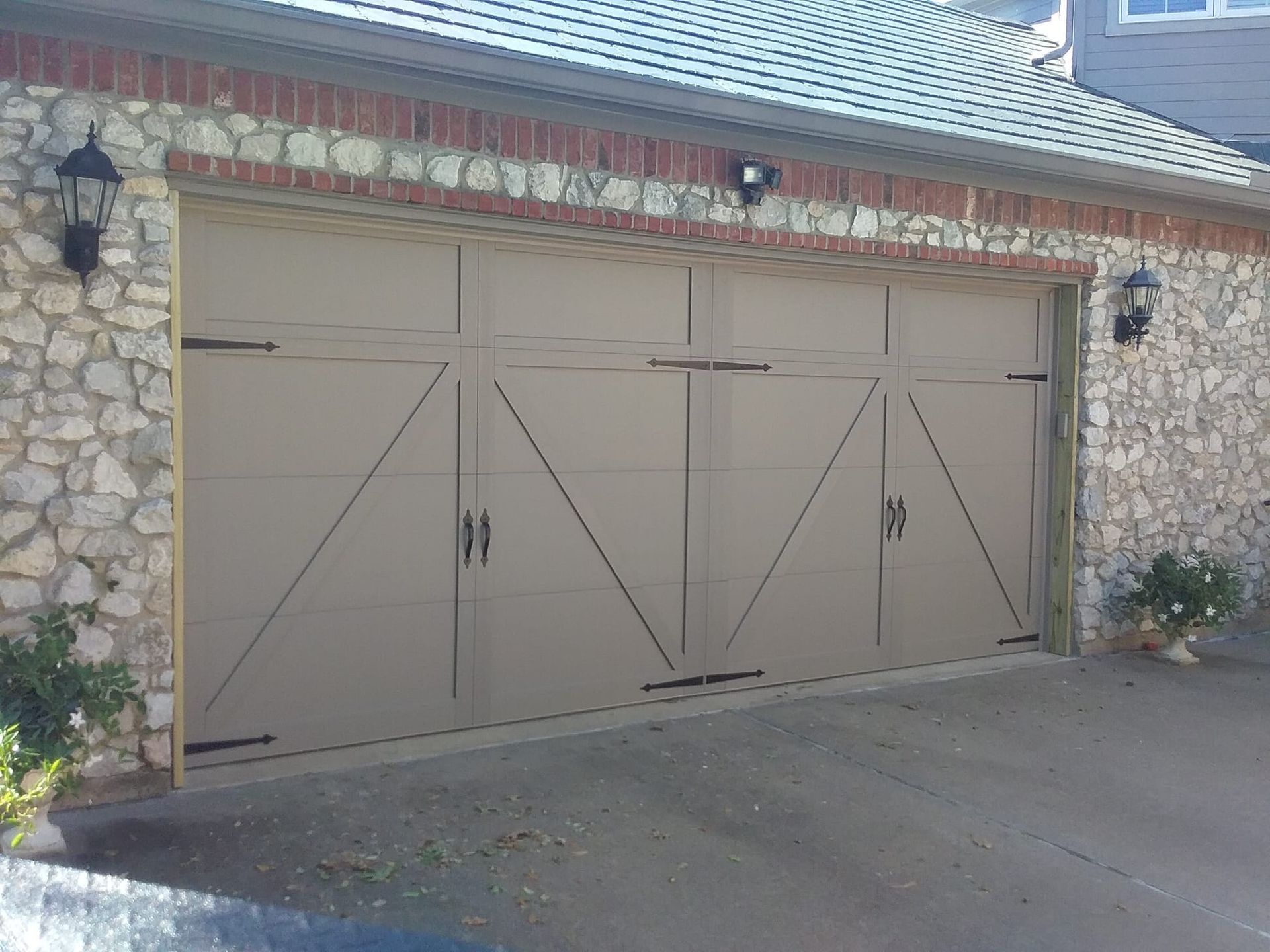 A tan, carriage-style double garage door with black hardware, set in a stone wall with brick trim and exterior lights.