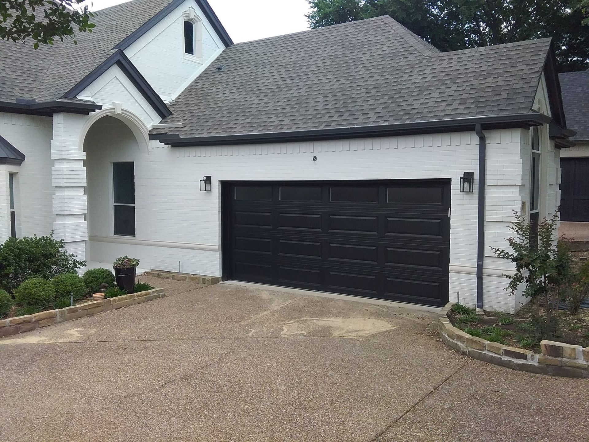 A white brick house with a dark garage door, a gray shingled roof, and a gravel driveway.