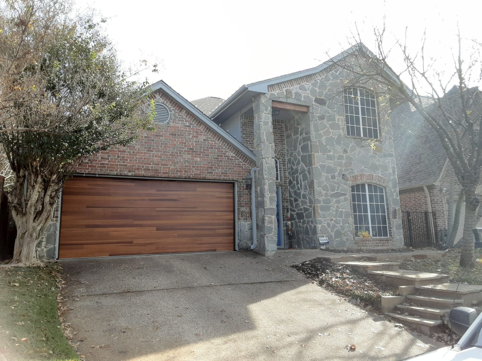 A two-story stone house with a wooden garage door and a gravel driveway in front of a tree.