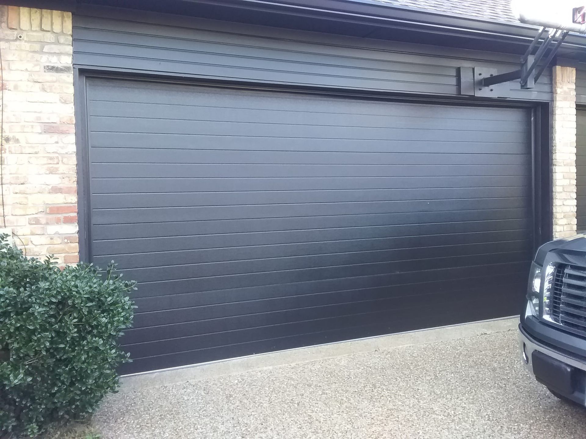 A modern black garage door with horizontal slats installed on a brick house, next to part of a dark SUV.