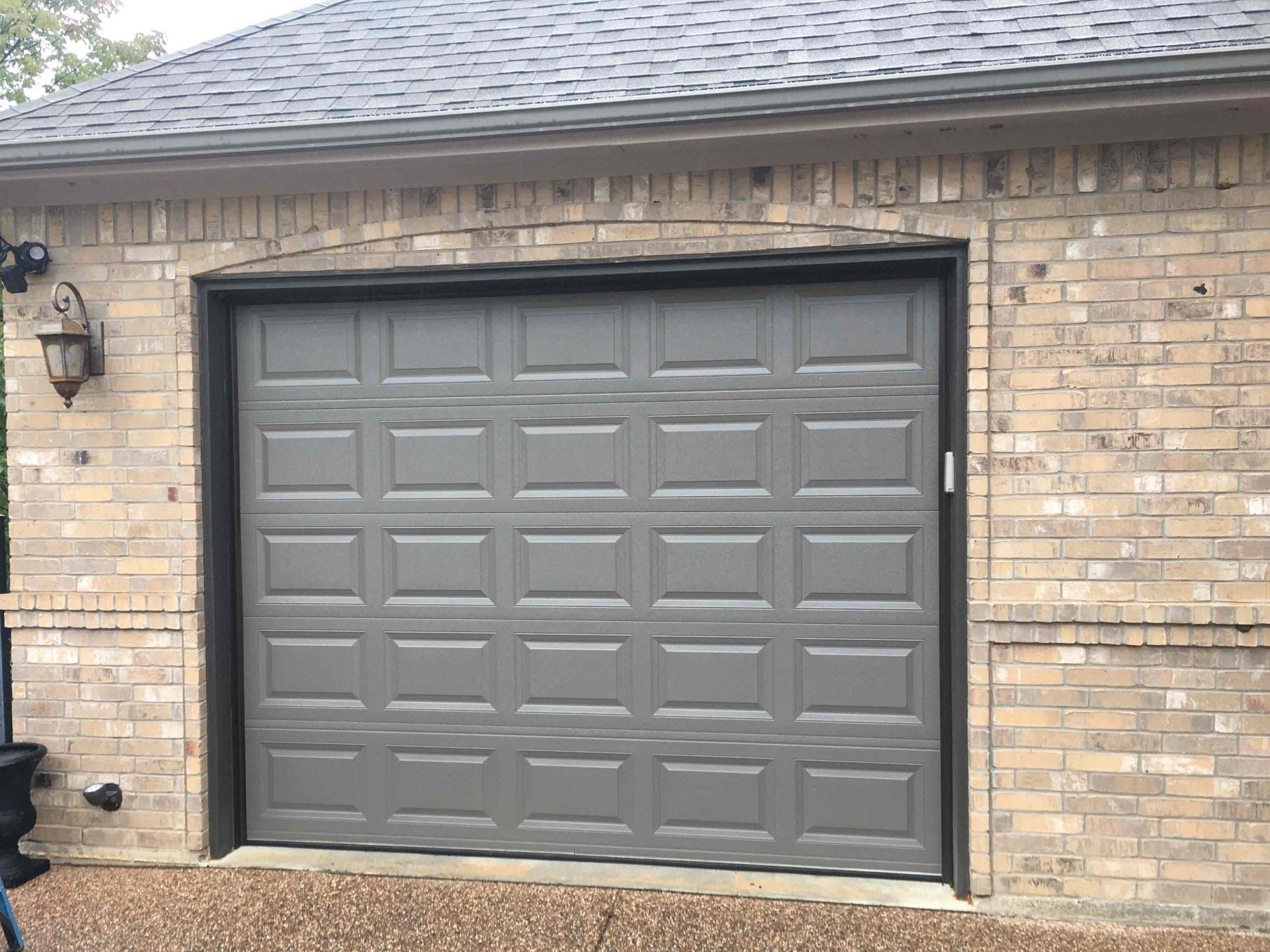 Gray garage door with a rectangular panel pattern installed in a tan brick building.
