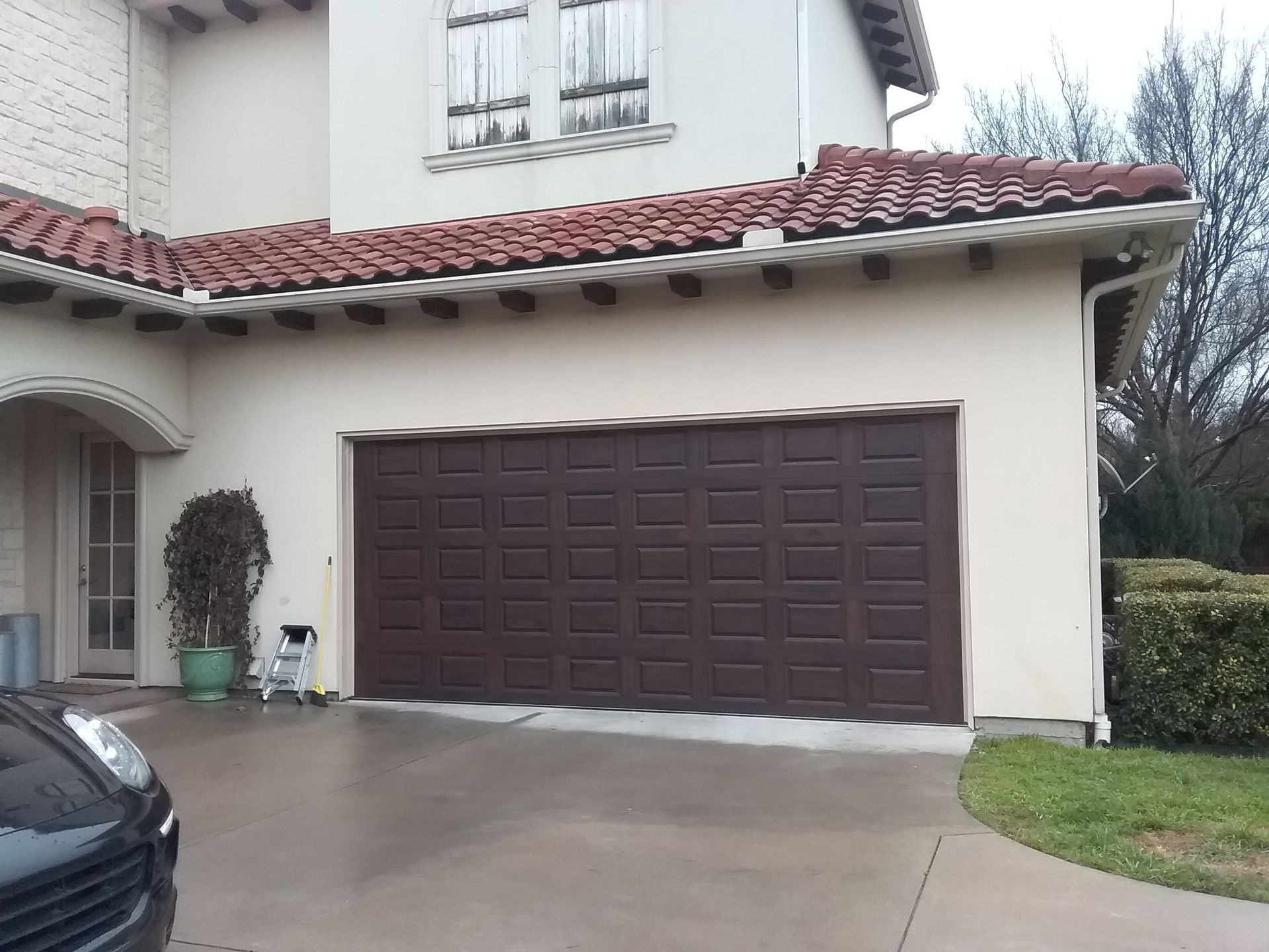 A house with a dark brown, multi-panel garage door, cream-colored stucco walls, and a reddish-brown tiled roof.