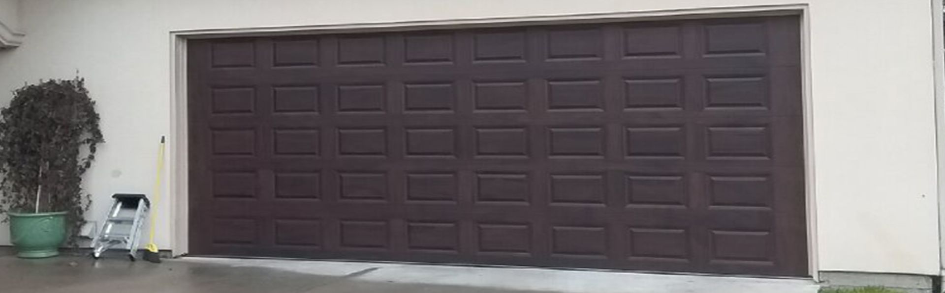 A dark brown, rectangular garage door with multiple recessed panels, set in a light-colored exterior wall.