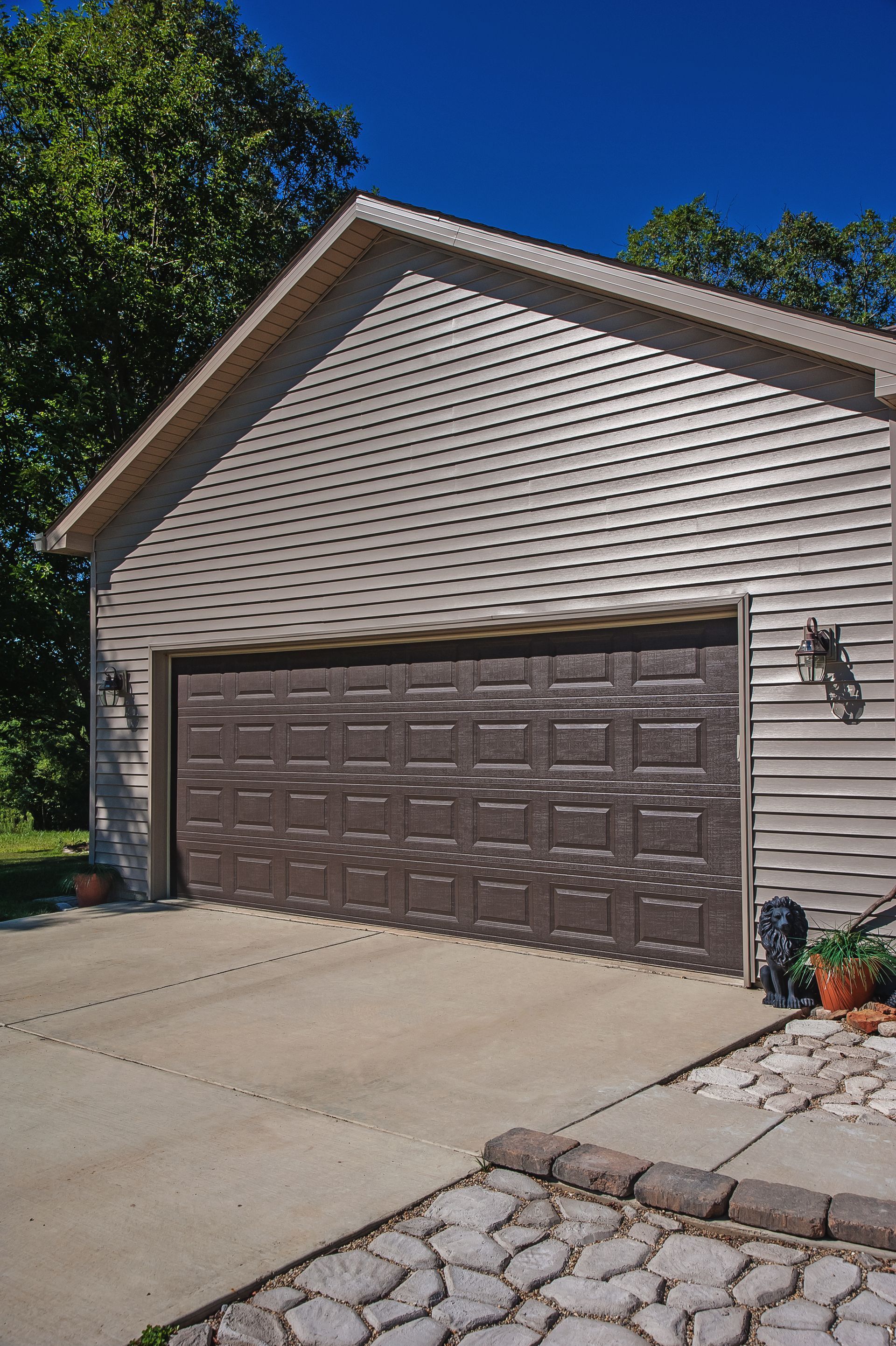 A detached garage with beige vinyl siding and a dark brown multi-panel door, situated under a bright blue sky.