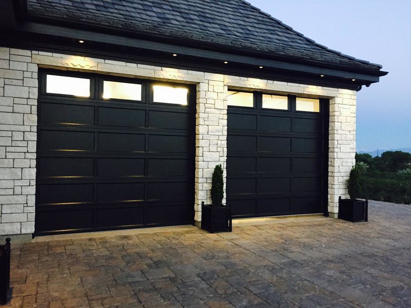 Two black garage doors with top row windows, framed by white stone masonry and set in a paved driveway at dusk.