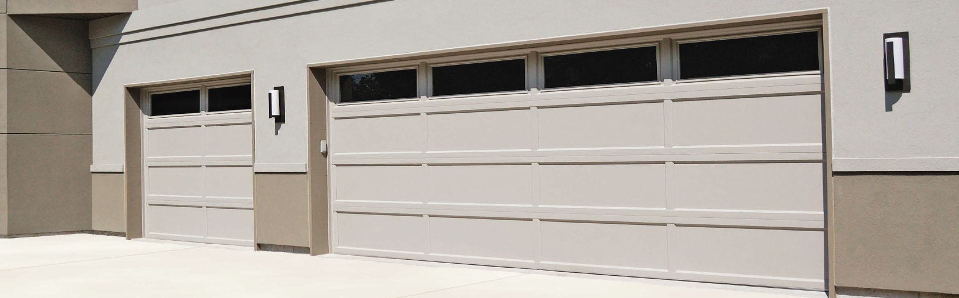 Two beige garage doors with rectangular windows at the top, installed in a light-colored stucco building exterior.