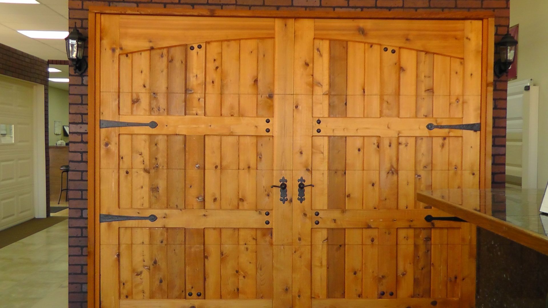 Double wooden carriage-style doors with black decorative hinges and handles set in a faux brick wall.