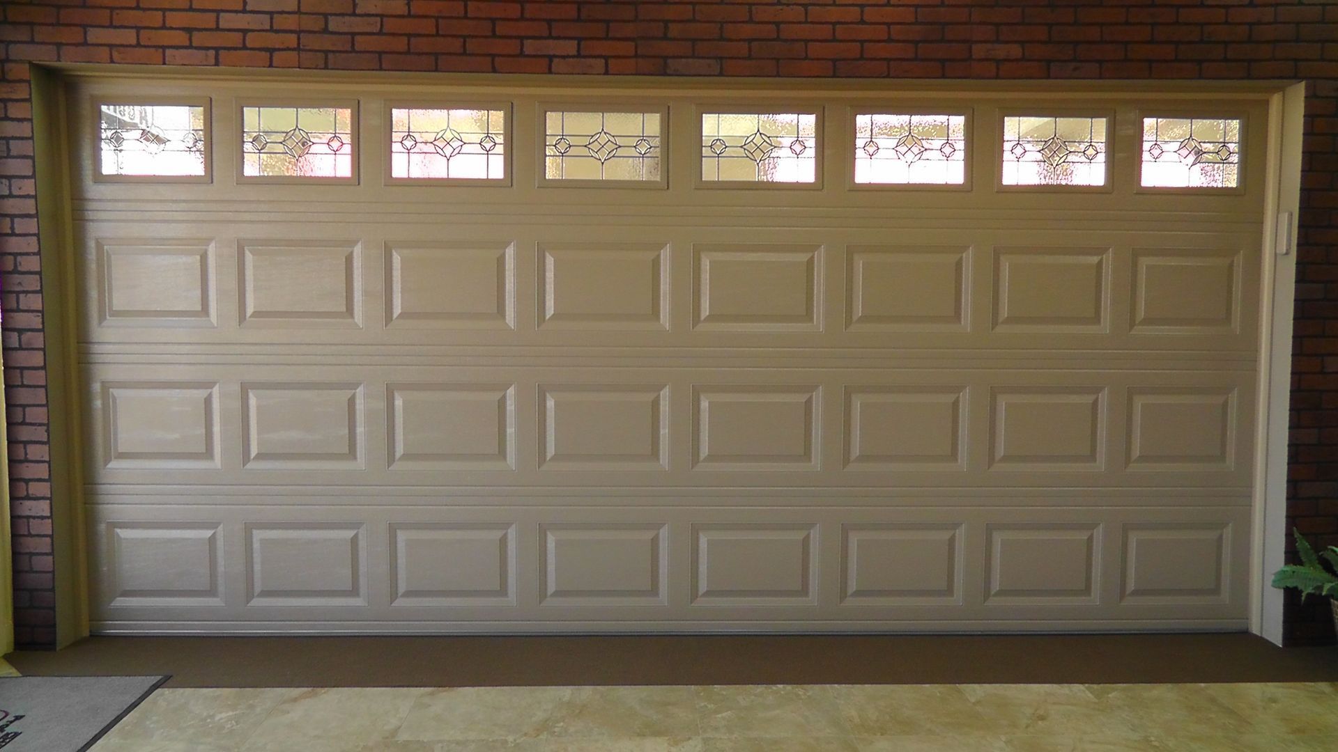 Tan garage door with four rows of panels and a top row of eight rectangular windows set into a brick wall.