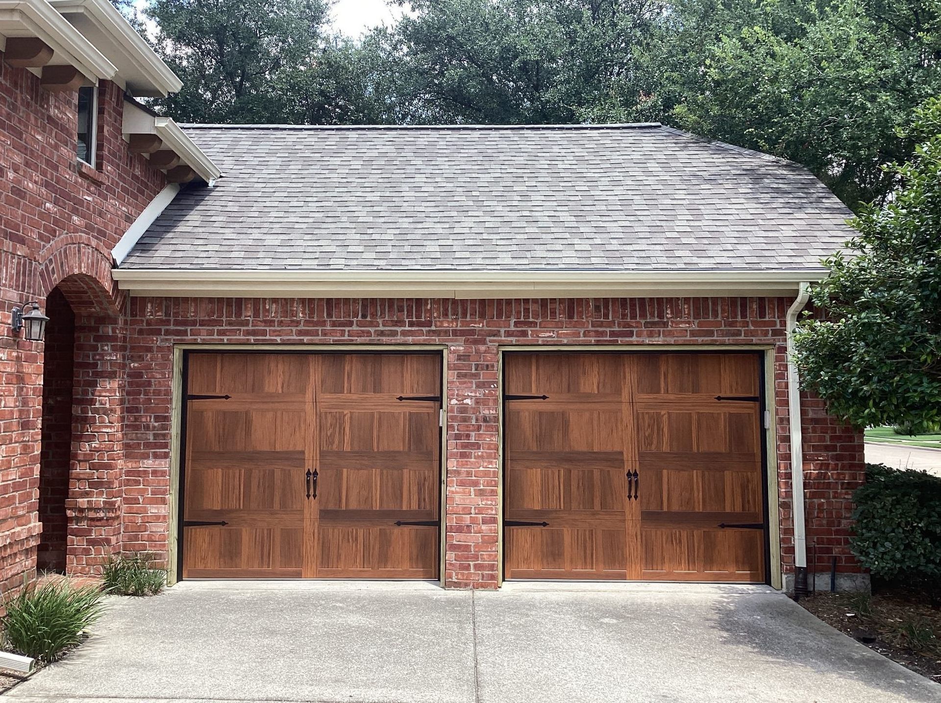 A two-car garage with wood-look carriage-style doors and dark hardware, attached to a red brick house exterior.