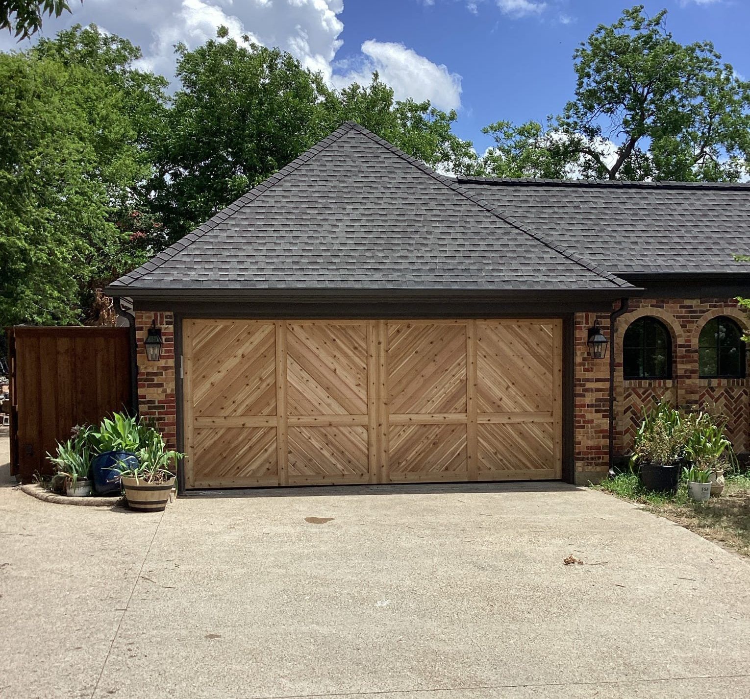 A two-car garage with light wood, diagonal-patterned doors, a dark shingled roof, brick walls, and potted plants outside.