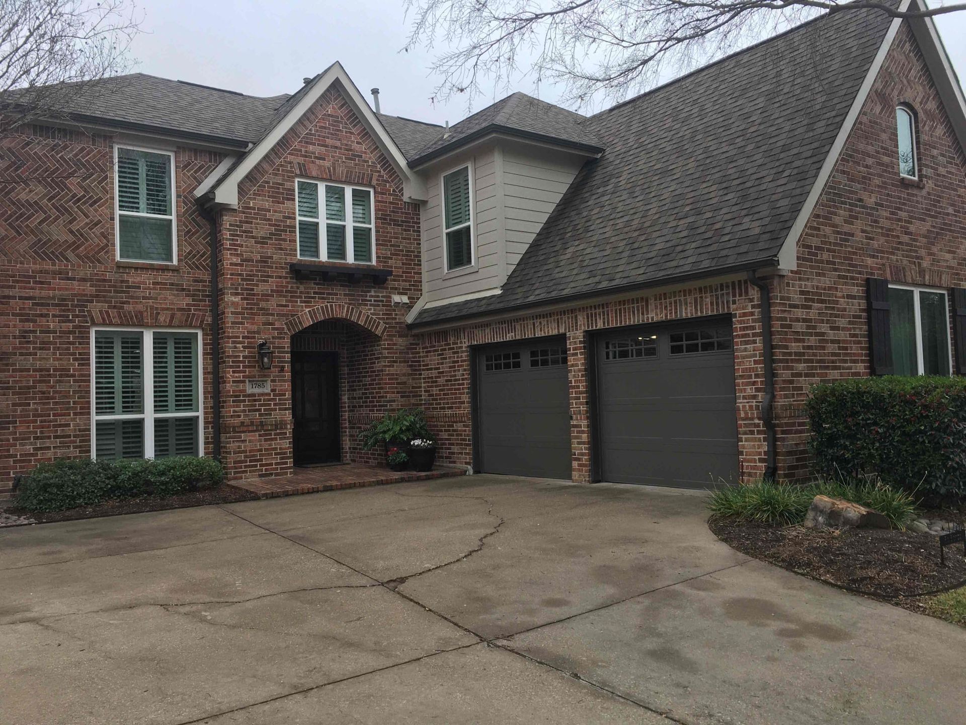 A two-story, red brick residential home with a grey shingled roof, two garage doors, and a paved driveway.