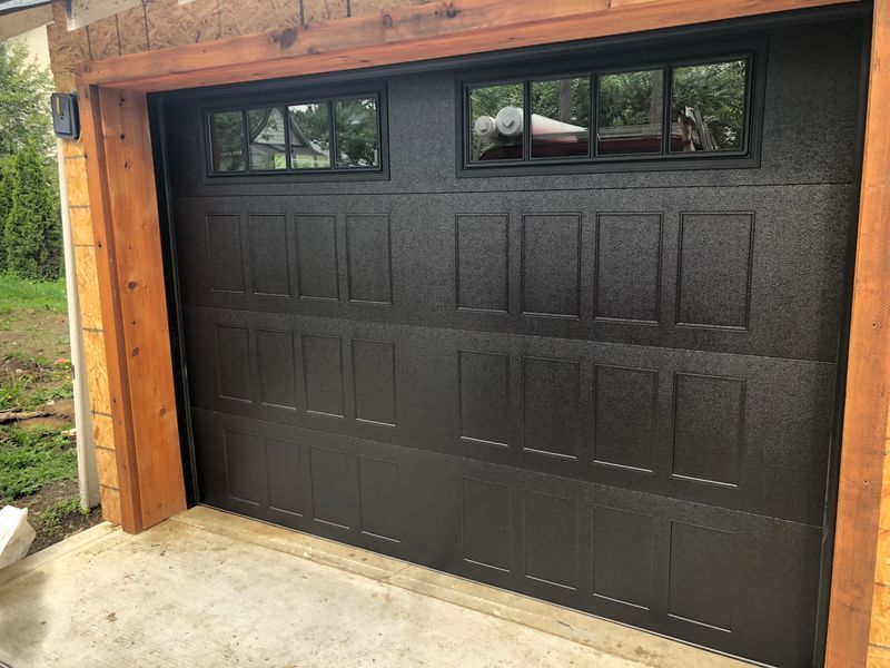 A black, paneled garage door with top row windows framed by natural wood trim, installed on a concrete driveway.