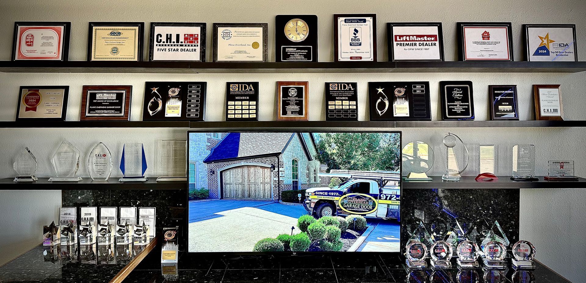Wall displays framed awards above a shelf with crystal trophies and a TV showing a truck.