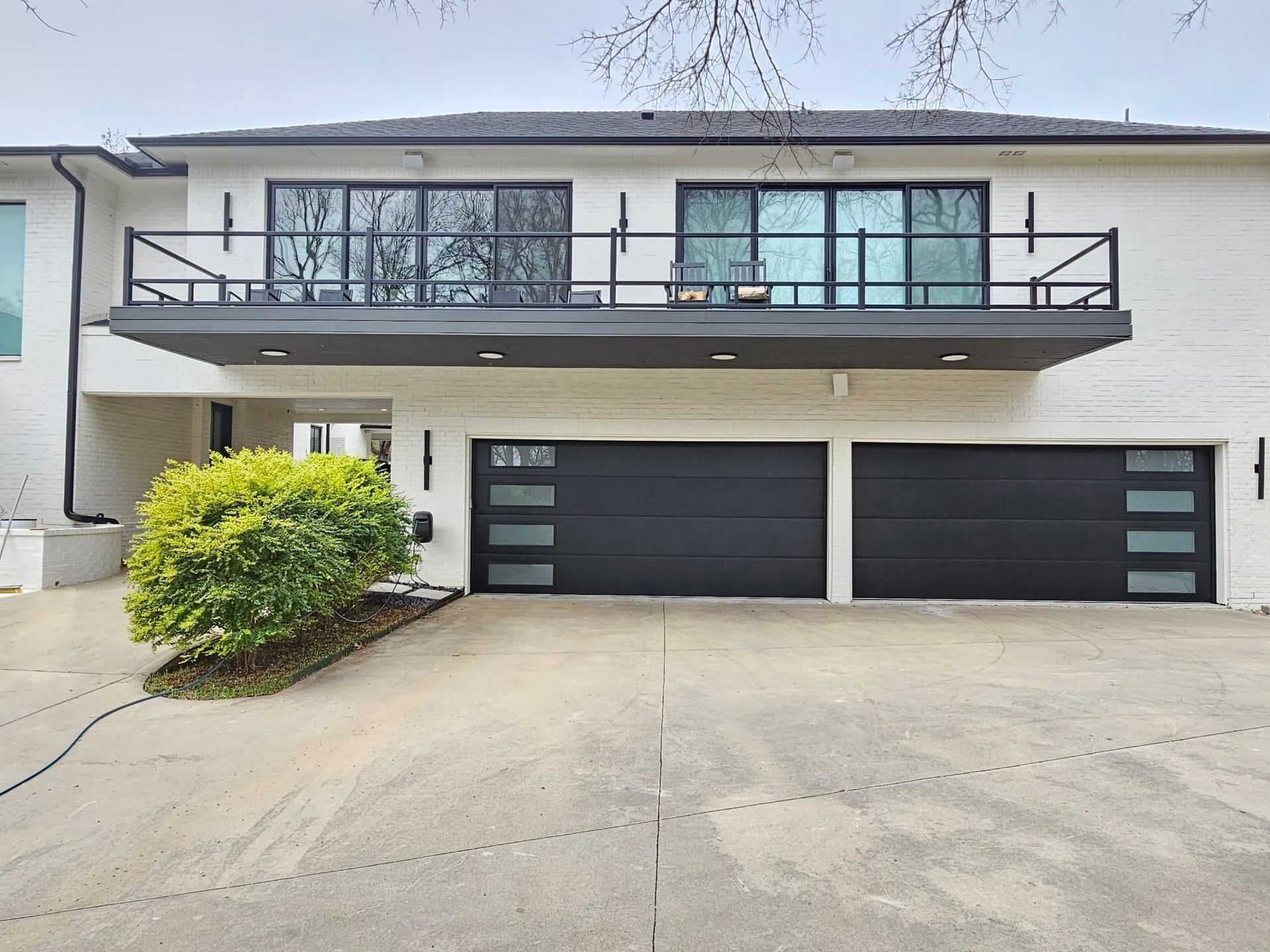 A two-story white house with two dark gray garage doors, a large balcony above them, and a green bush in the driveway.
