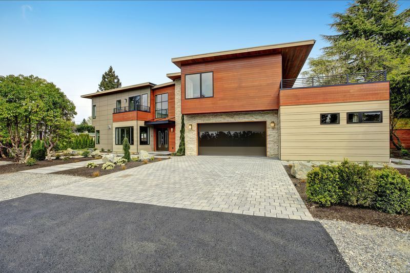 Modern two-story house with a flat roof, wood siding, stone accents, and a paved driveway under a blue sky.