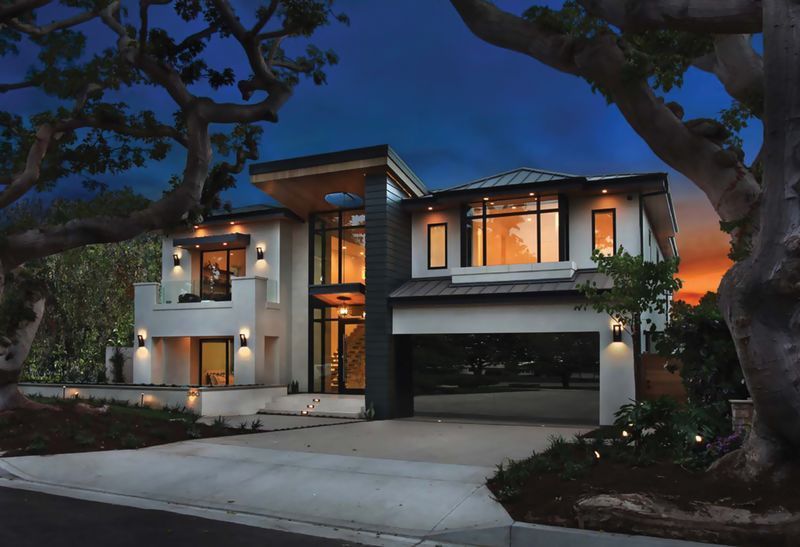 A modern two-story home at dusk, featuring a driveway, illuminated windows, and surrounding trees against a sunset sky.