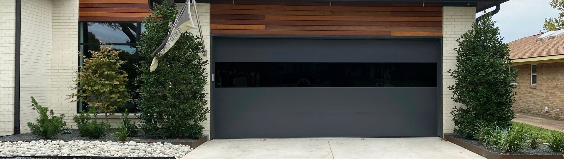 Modern gray garage door with dark horizontal window, flanked by green bushes and white brick facade. Wooden overhang.