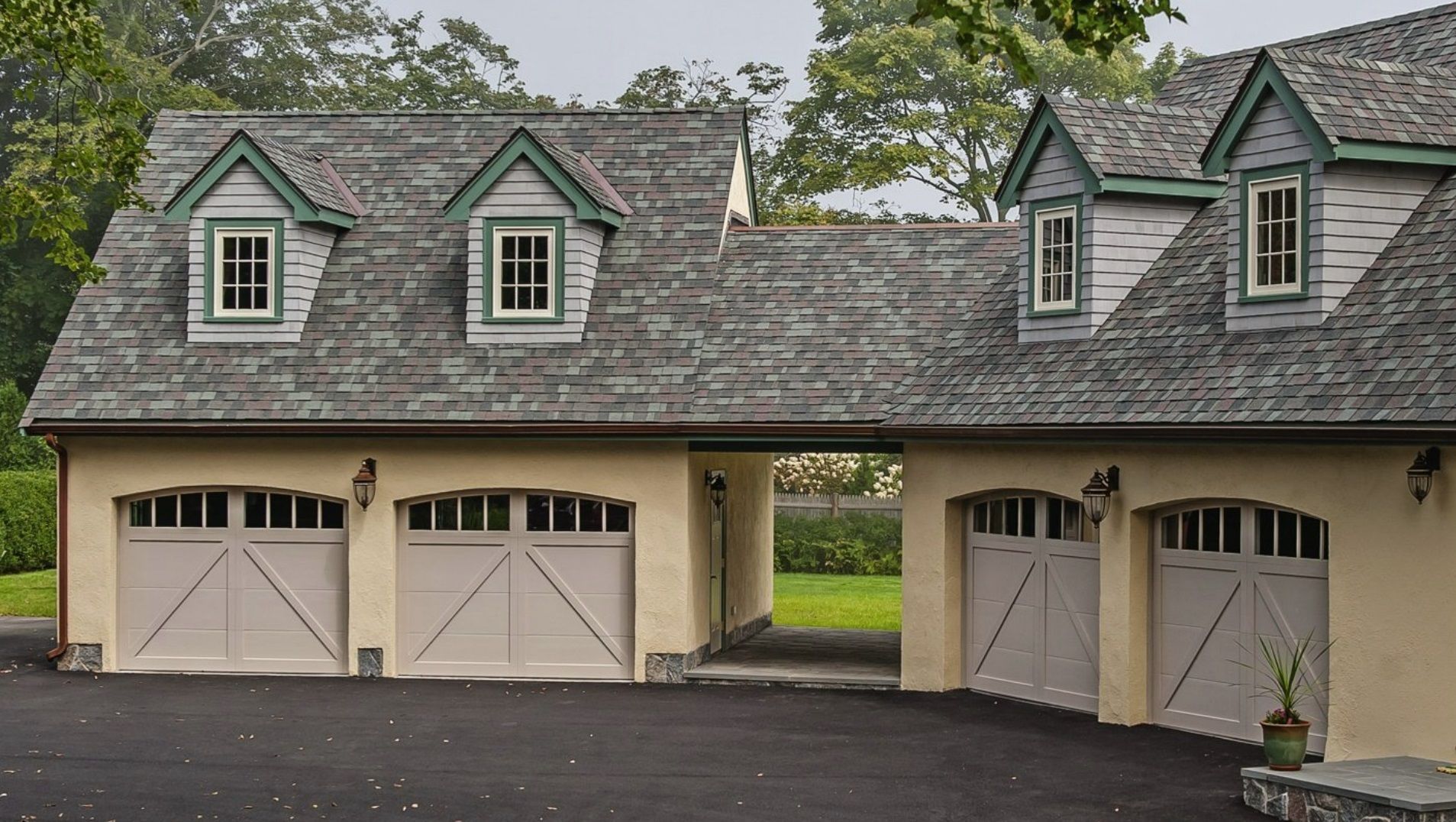 A tan, four-car carriage house with a shingled roof, arched garage doors, and four dormer windows, set in a green landscape.