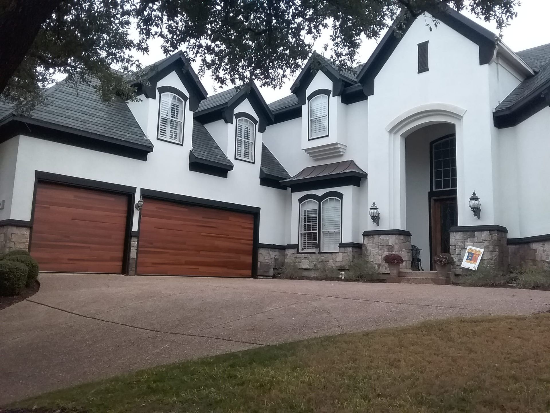 A two-story white house with dark trim, wood-toned garage doors, a stone base, and a paved driveway.