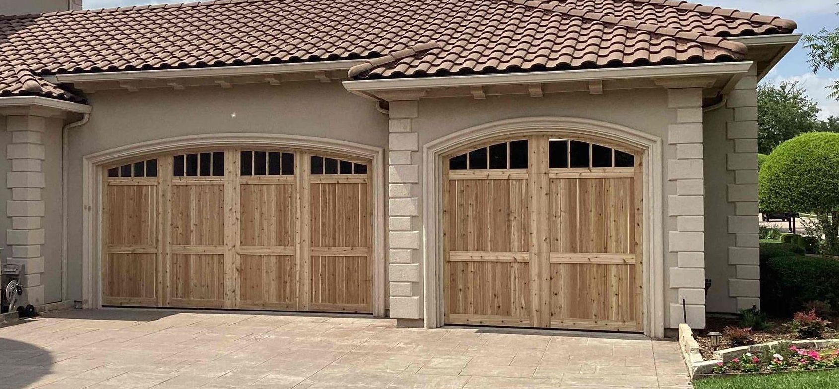 Two wooden garage doors with arched tops, windows, and decorative trim on a beige stucco house with a tile roof.