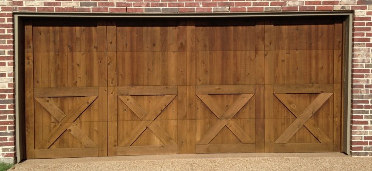 A wide wooden garage door with an X-shaped farmhouse design, set into a red brick exterior wall.