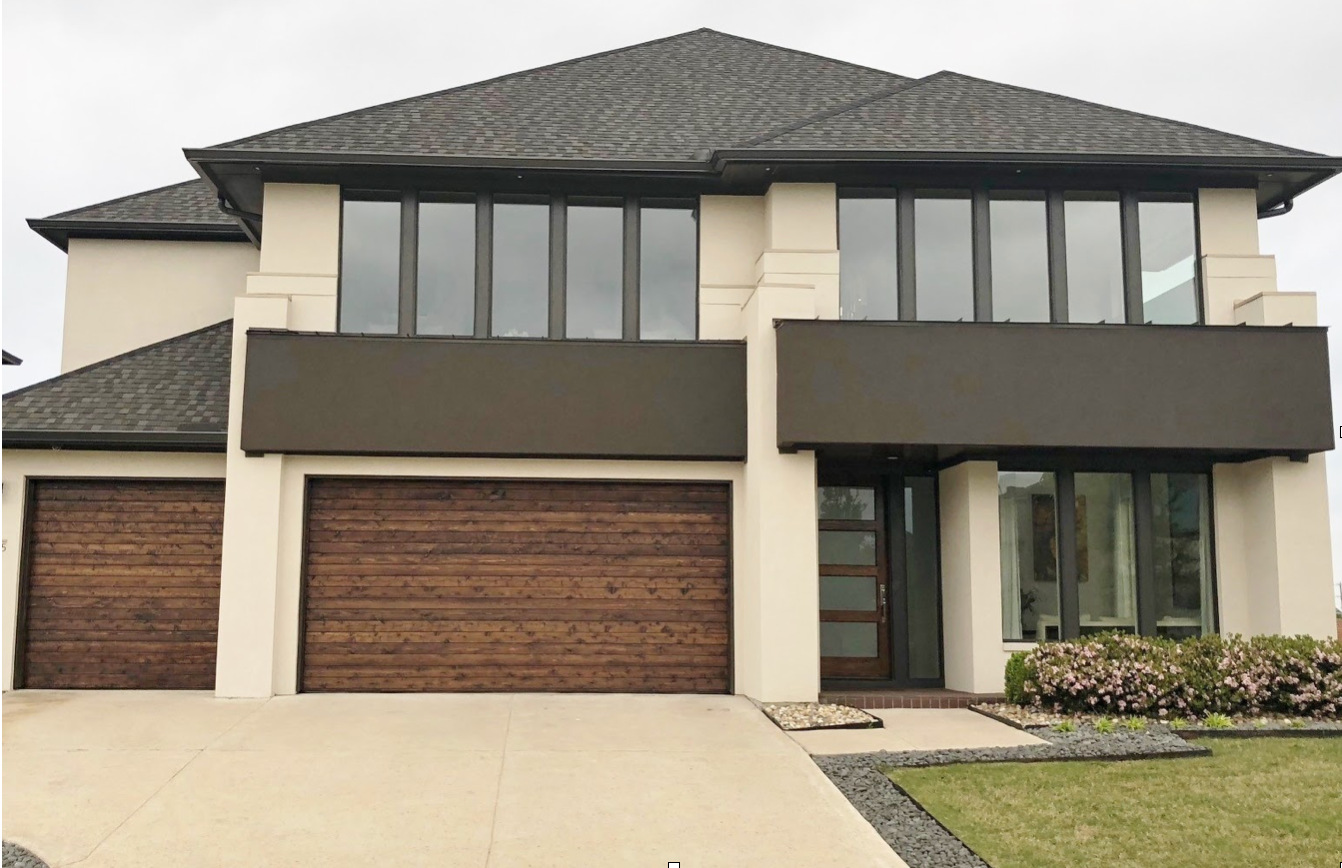 A two-story house with a beige stucco exterior, a dark gray roof, wood-toned garage doors, and modern, dark-framed windows.