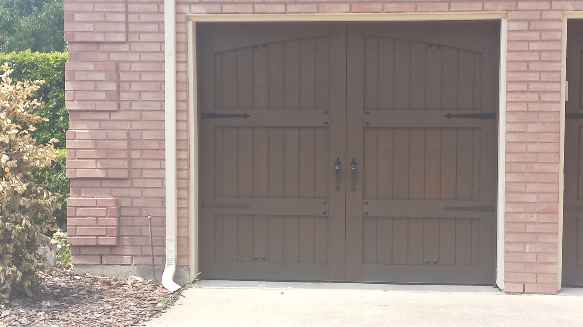A brown, carriage-style garage door with decorative hinges and handles set in a red brick wall.