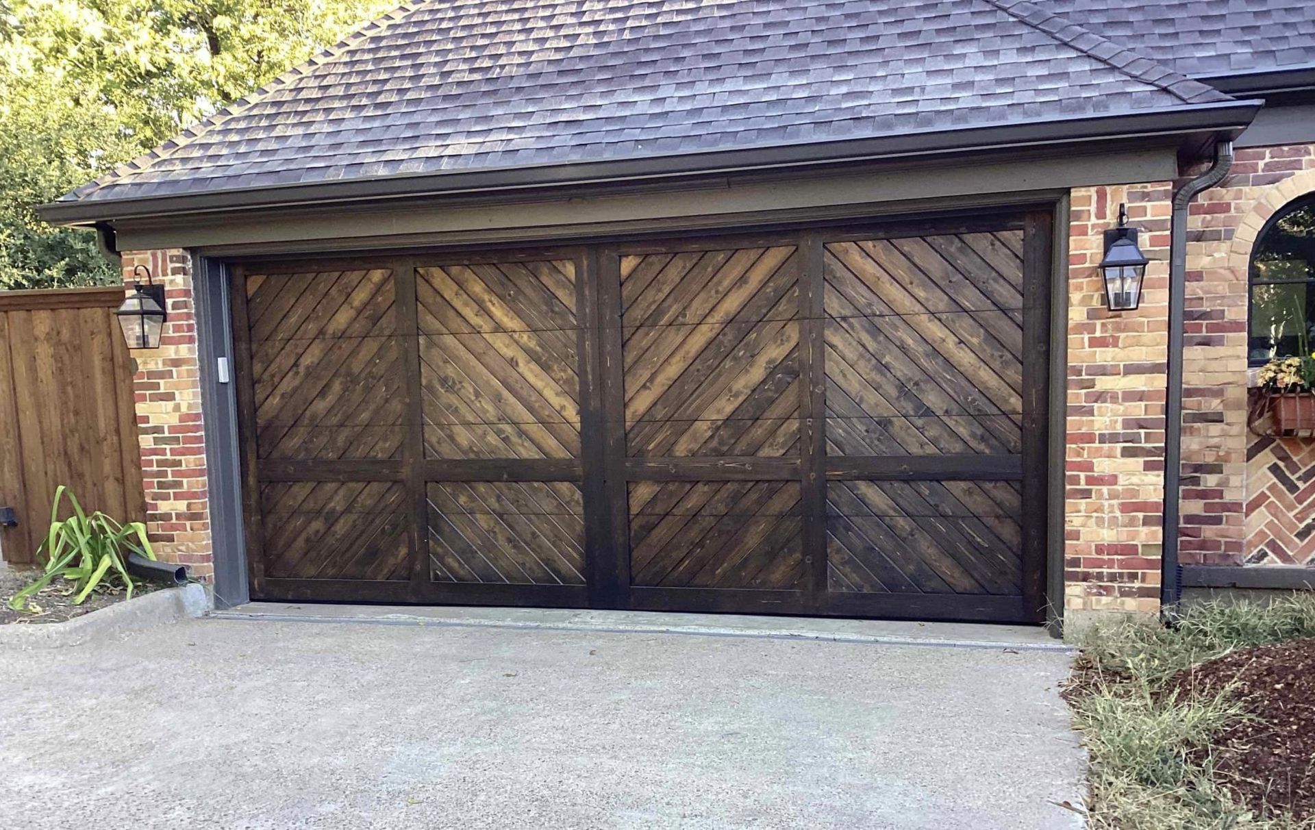 A wide, wooden garage door featuring a dark, diagonal herringbone pattern on a brick house exterior.