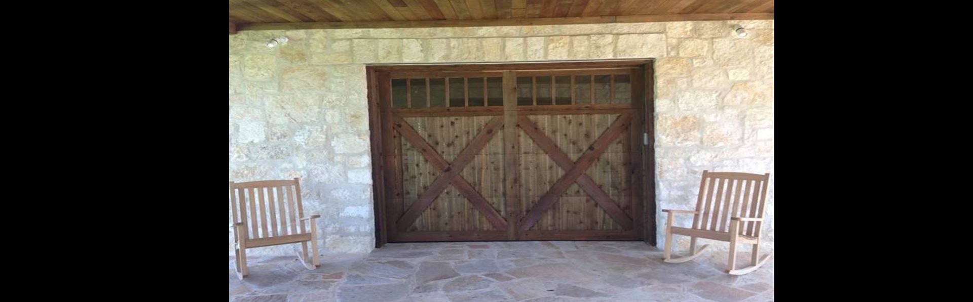 Two wooden rocking chairs on a stone patio in front of a rustic garage door with an X-brace design and small windows.