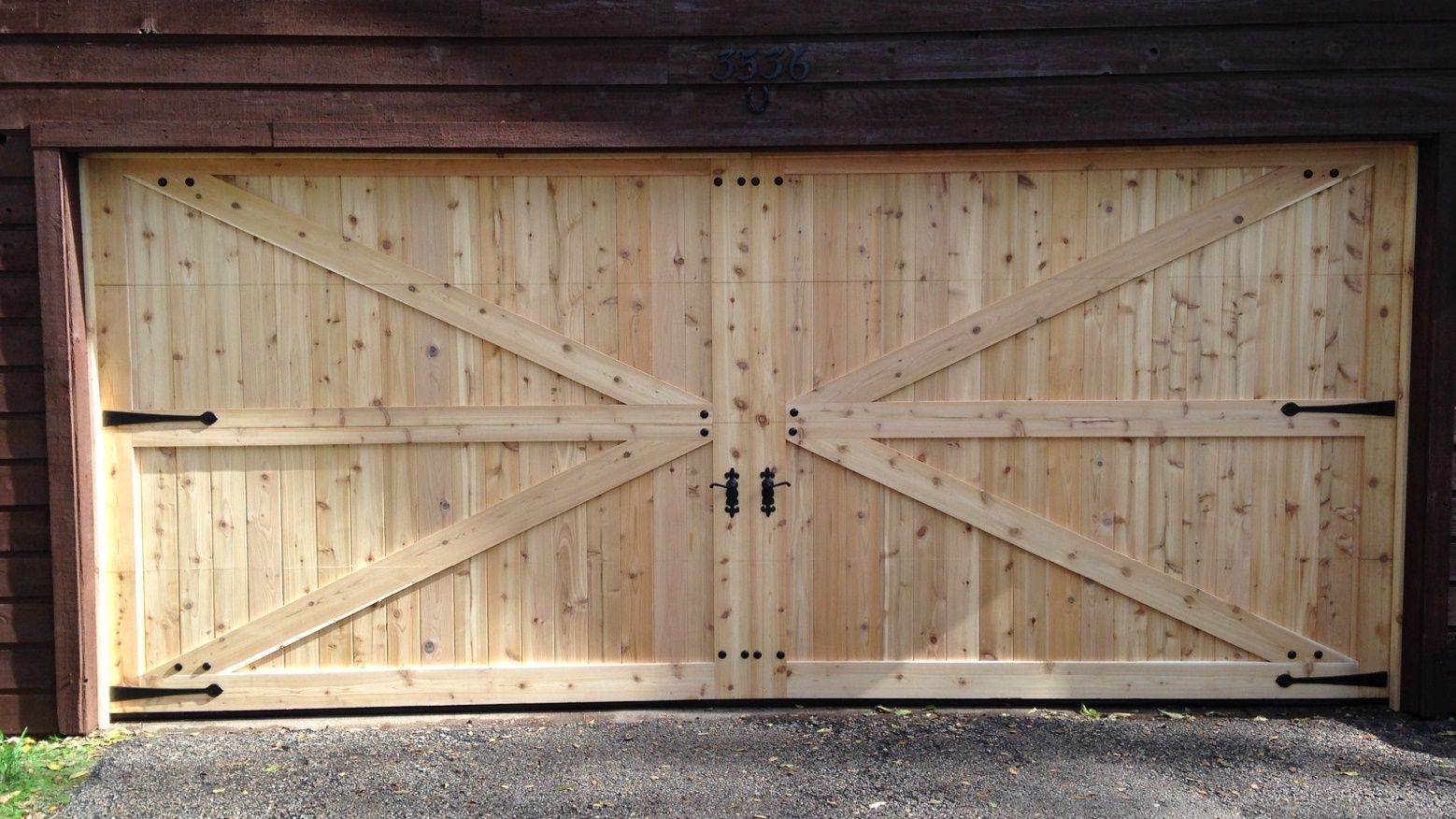 Double wooden garage doors made of light-colored, knotty pine with black metal hardware and a decorative X-brace design.