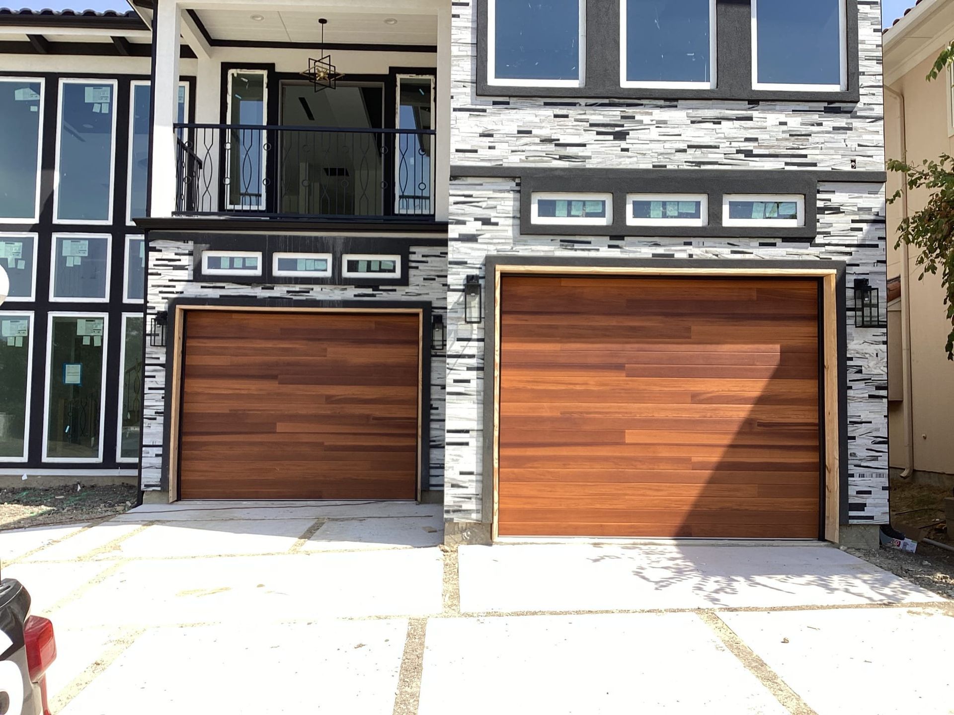 A modern two-story home exterior with two warm-toned wood garage doors, stone siding, and large glass windows.