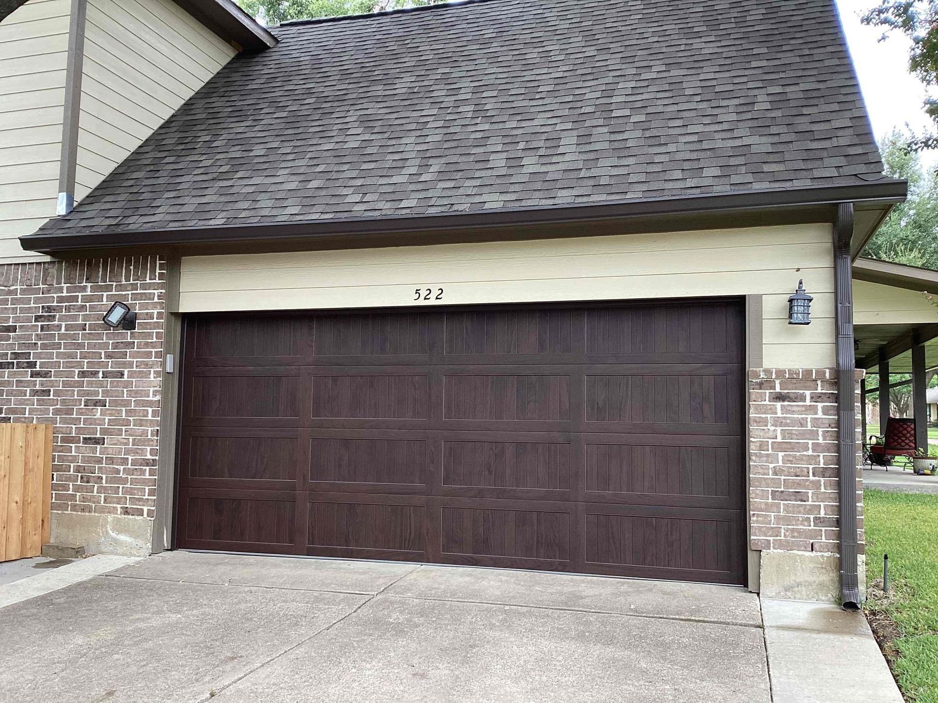 A dark brown garage door with multiple rectangular panels, framed by brick and light-colored siding on a house exterior.