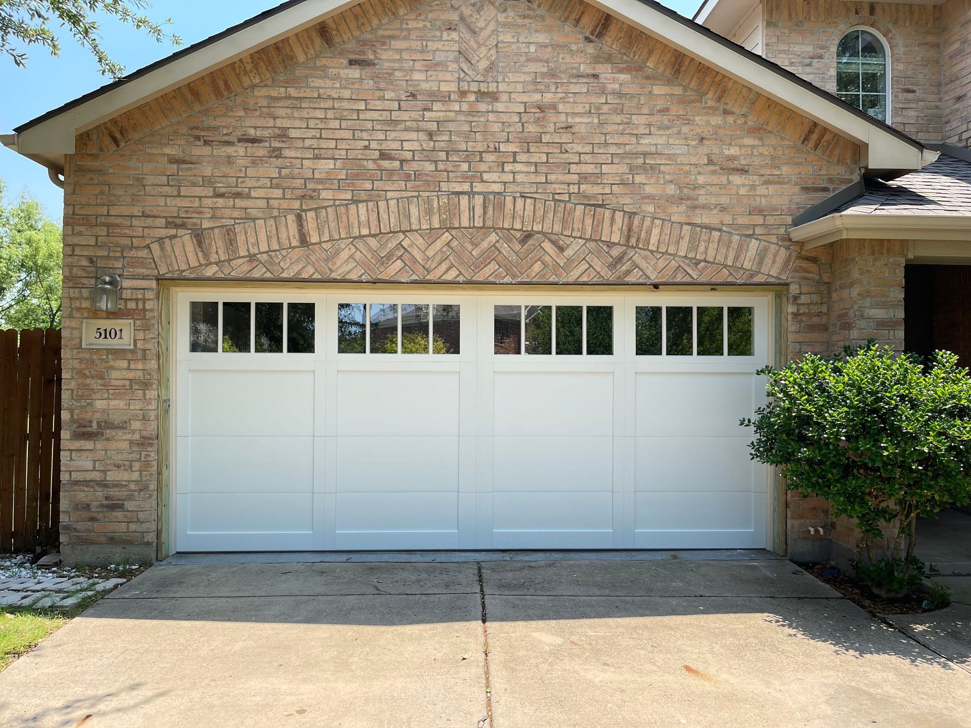 A wide, white four-panel garage door with rectangular windows under a brick archway on a suburban home.