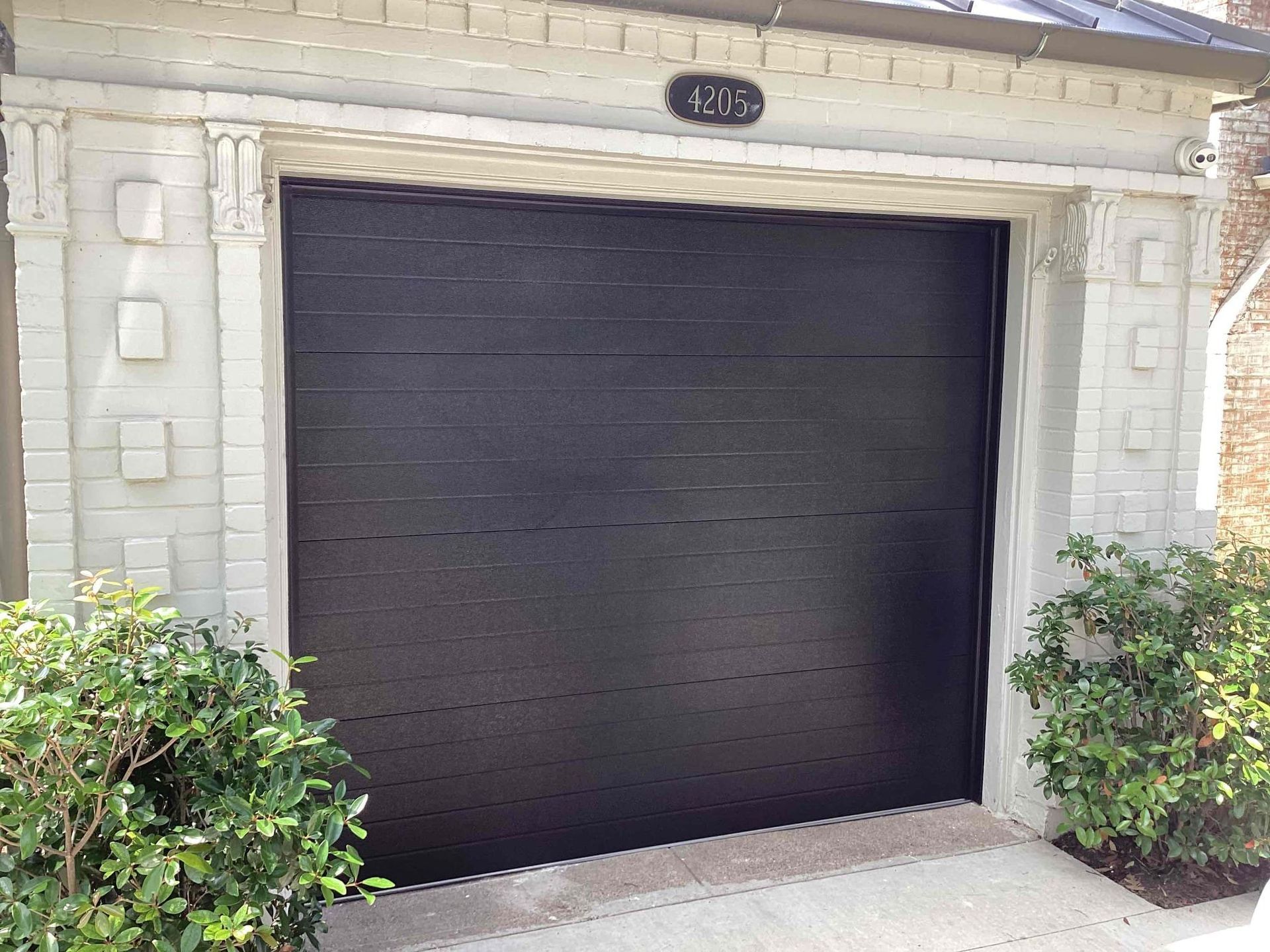 A black, horizontally paneled garage door set within a light-colored, decorative brick exterior frame with landscaping.