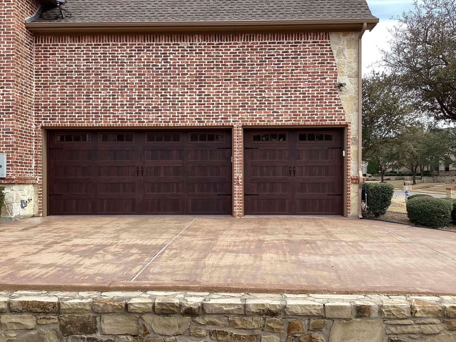 A two-car garage with dark brown carriage-style doors on a brick house, fronted by a concrete driveway and stone wall.