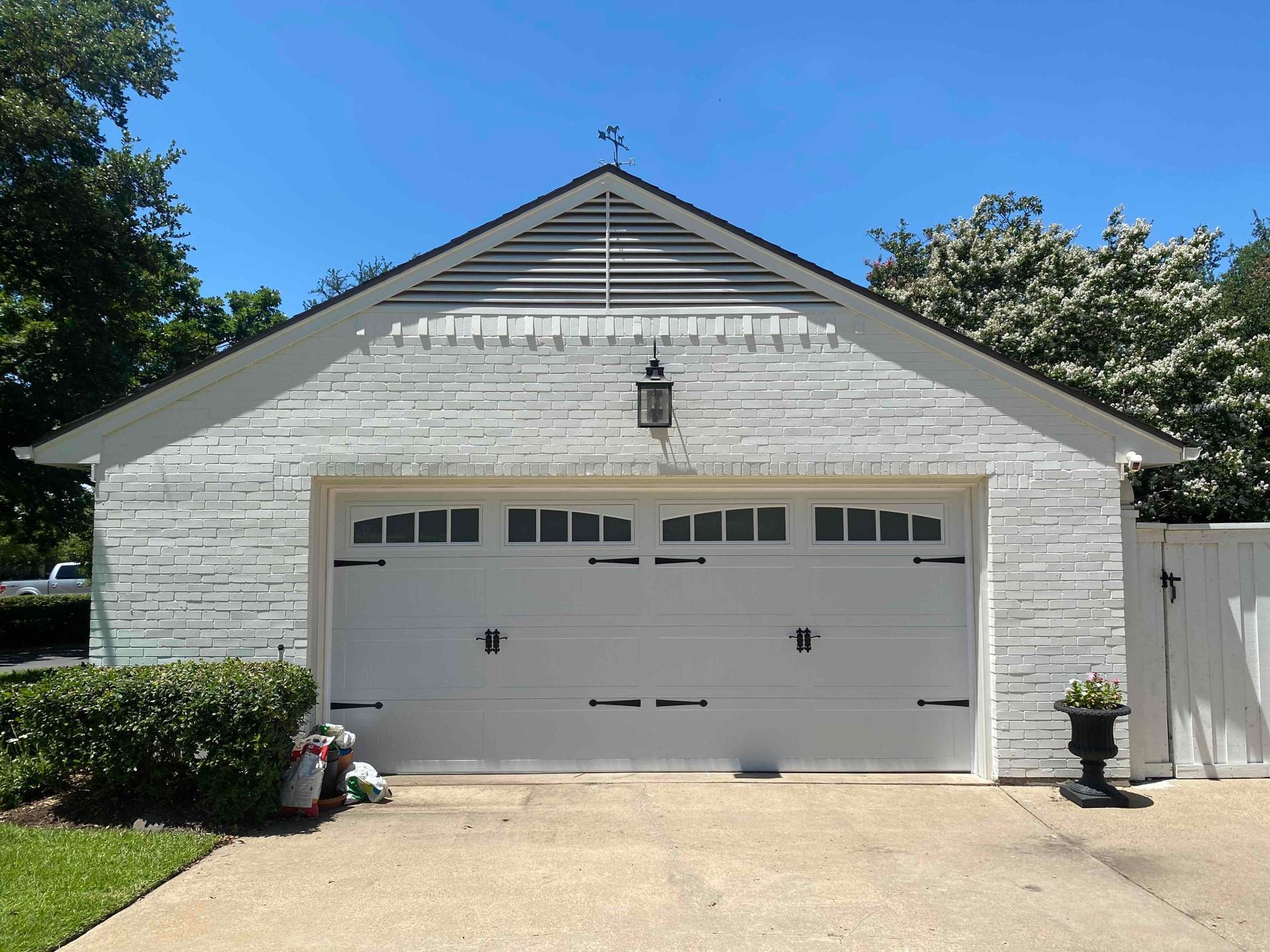 A white brick detached garage with a white sectional door featuring windows, a louvered vent, and a black driveway lamp.