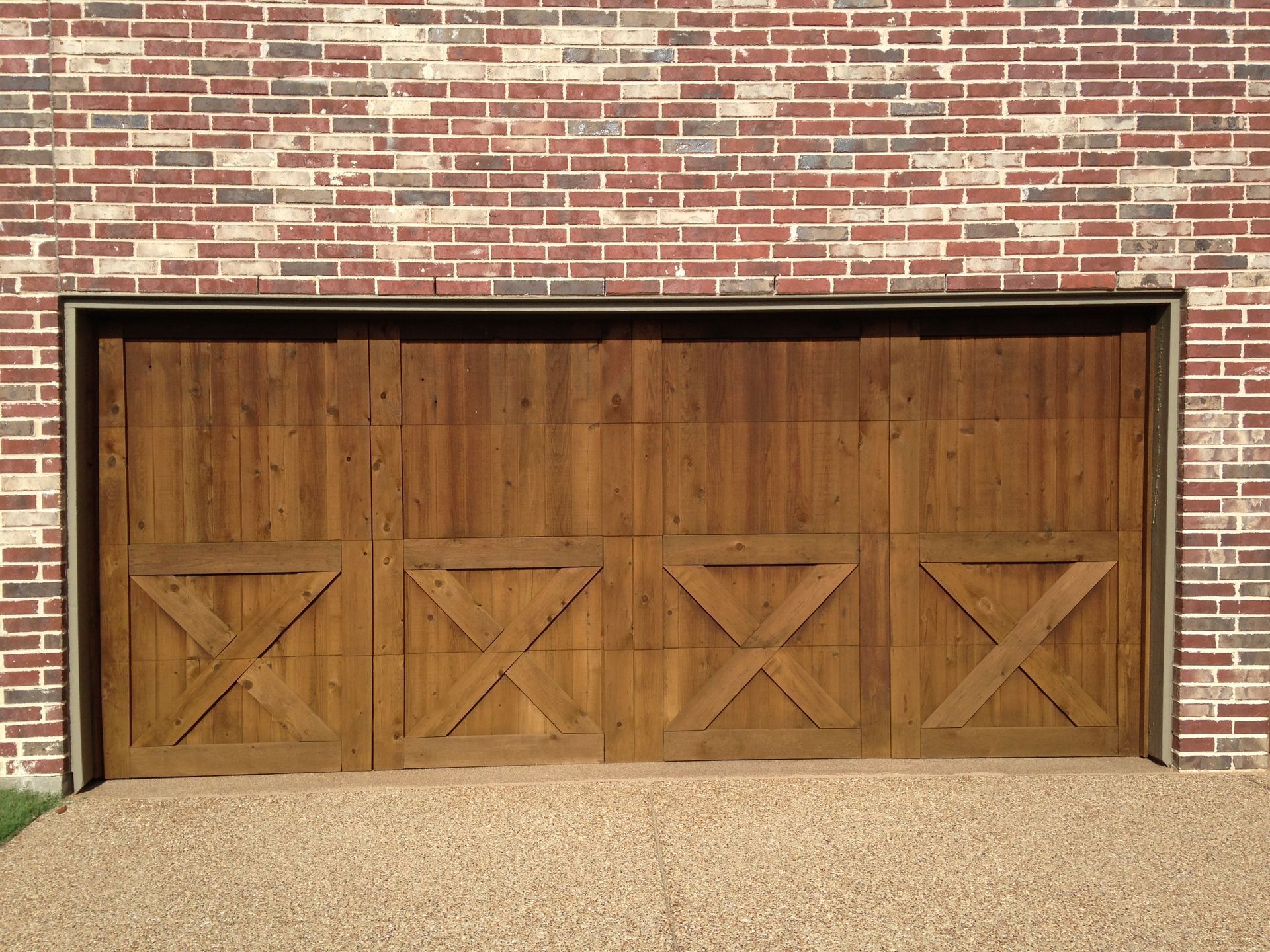Wooden garage door with X-brace designs installed in a multi-colored brick wall above a pebbled driveway.
