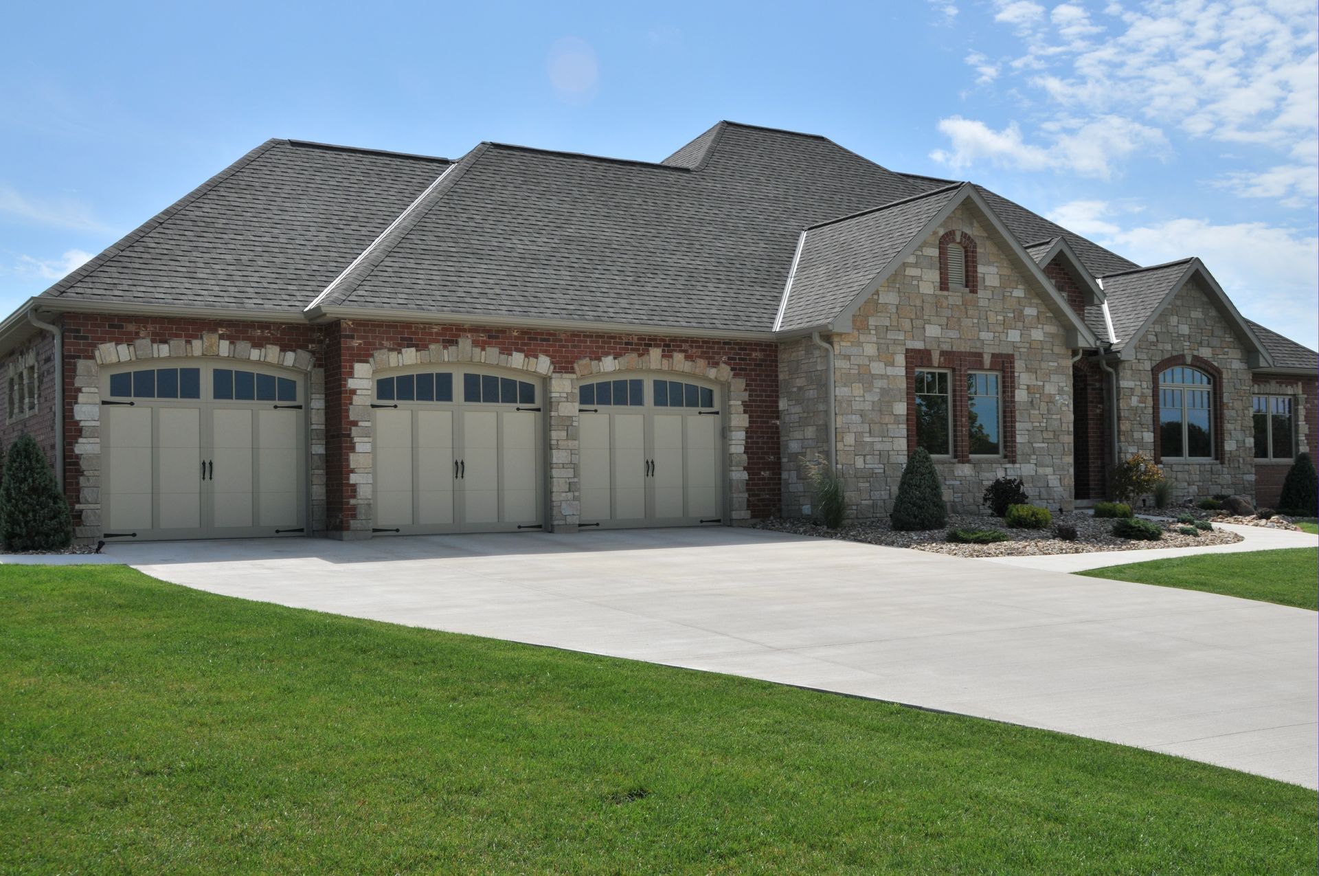 A stone and brick house with a three-car garage and a concrete driveway, set against a blue sky with light clouds.
