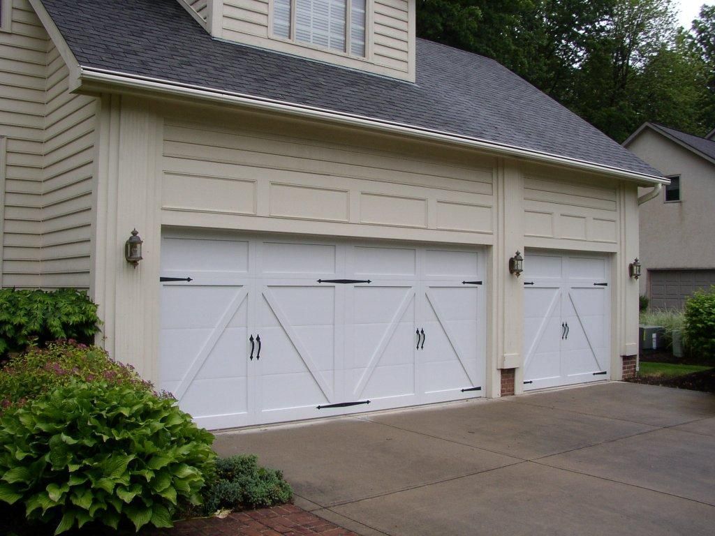 A cream-colored garage with three white carriage-style doors and dark metal hinges, surrounded by shrubs on a driveway.