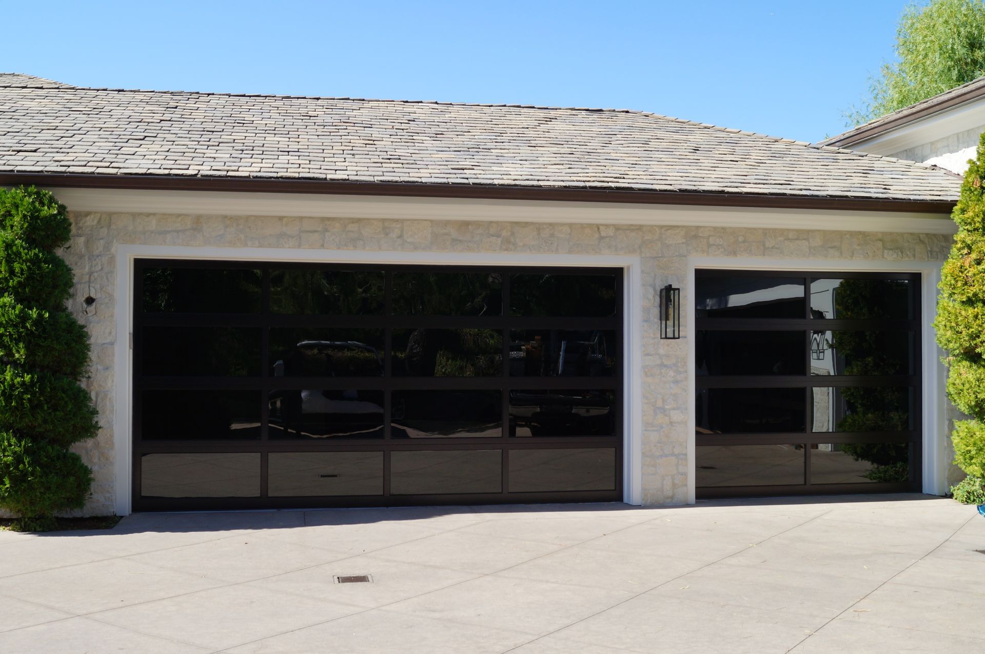 A modern home with two dark, multi-panel glass garage doors set in a stone exterior wall with green shrubs.