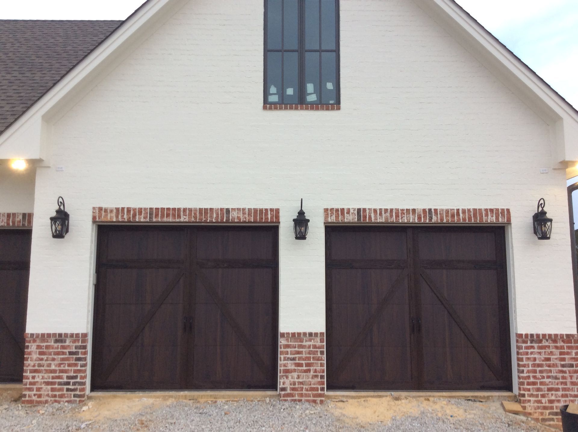 White brick exterior of a garage with two dark wooden carriage-style doors, brick trim, and outdoor wall lanterns.