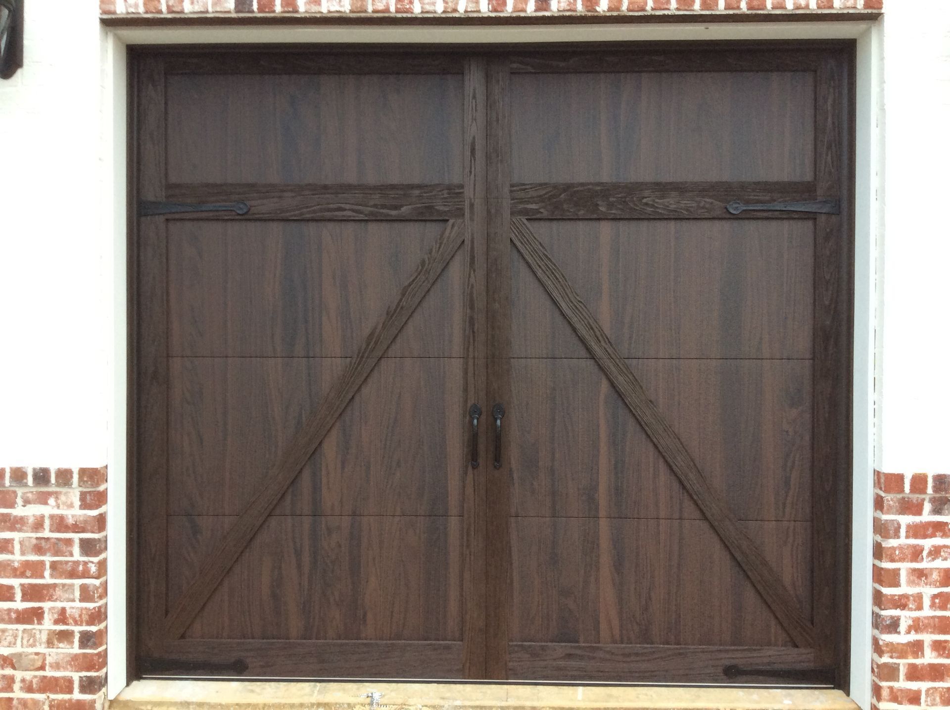 A pair of dark wood carriage-style garage doors with diagonal bracing and black hinges, framed by brick walls.