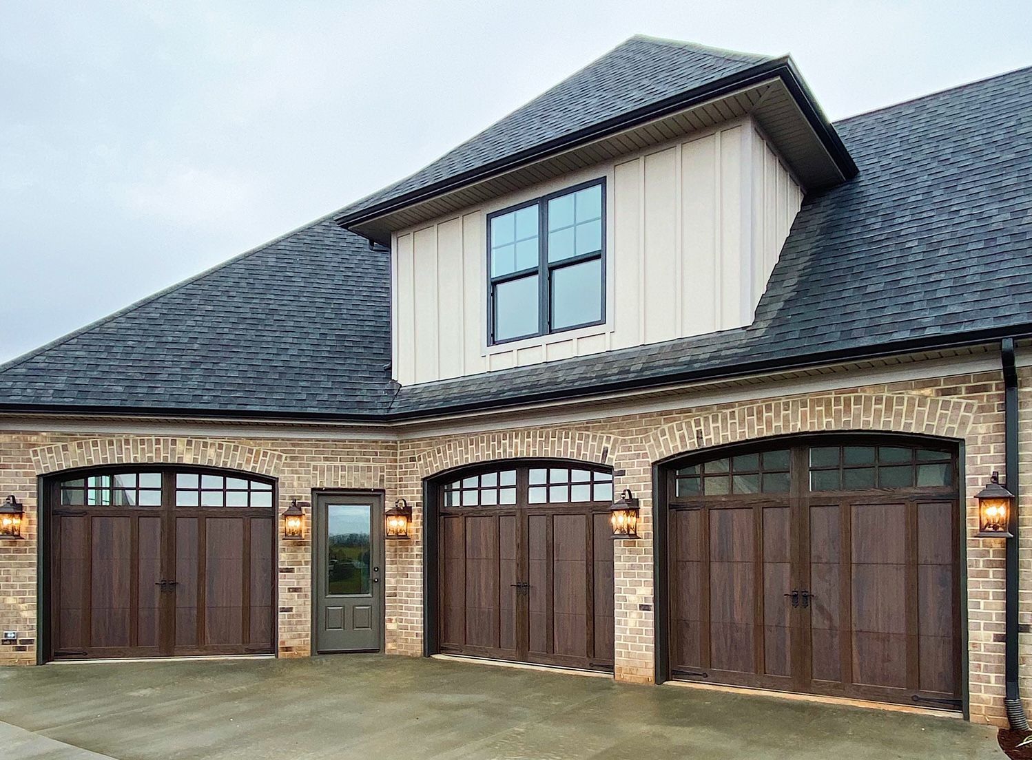 Three-car garage with arched wooden doors, a central door, and a dormer window above on a brick building with a dark roof.