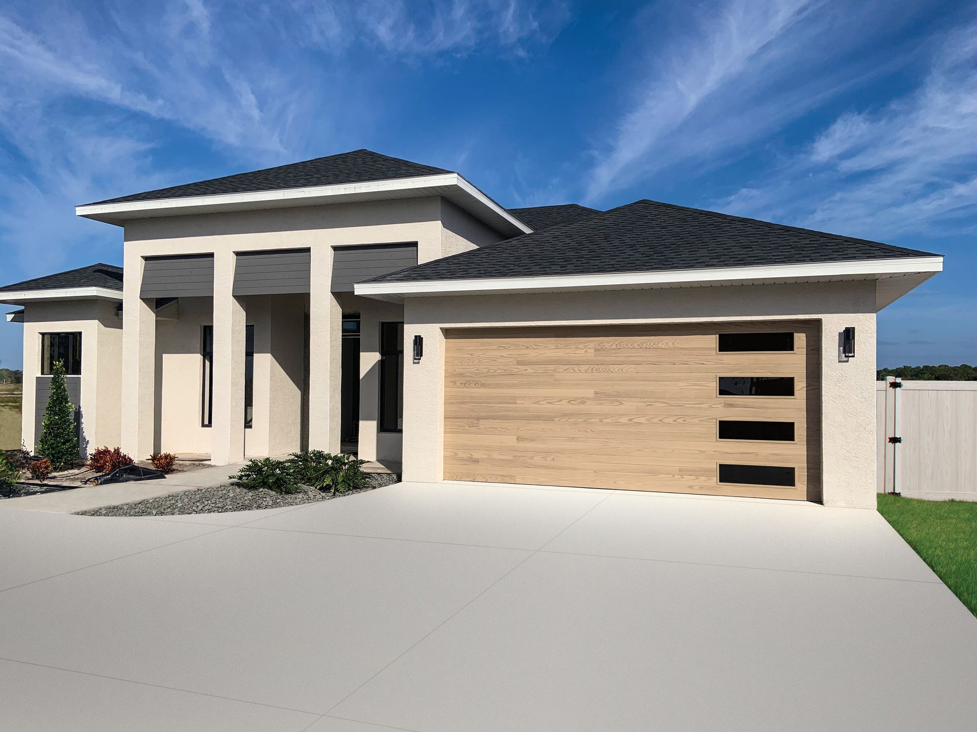 Single-story house with light beige stucco, a dark grey tiled roof, and a wood-toned garage door under a bright blue sky.