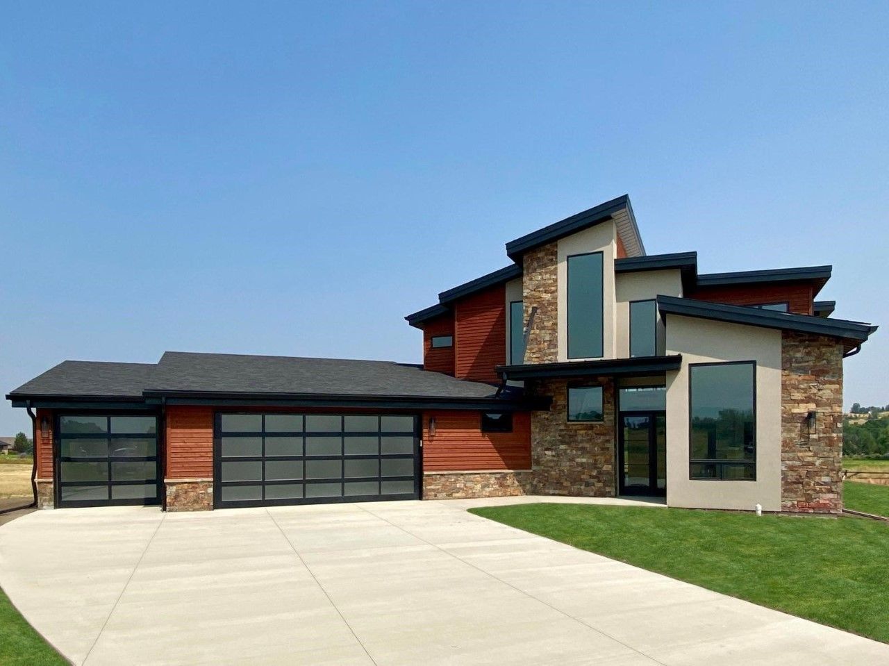 A modern two-story home with a tan stone facade, wood siding, large glass windows, and a three-car garage under a blue sky.