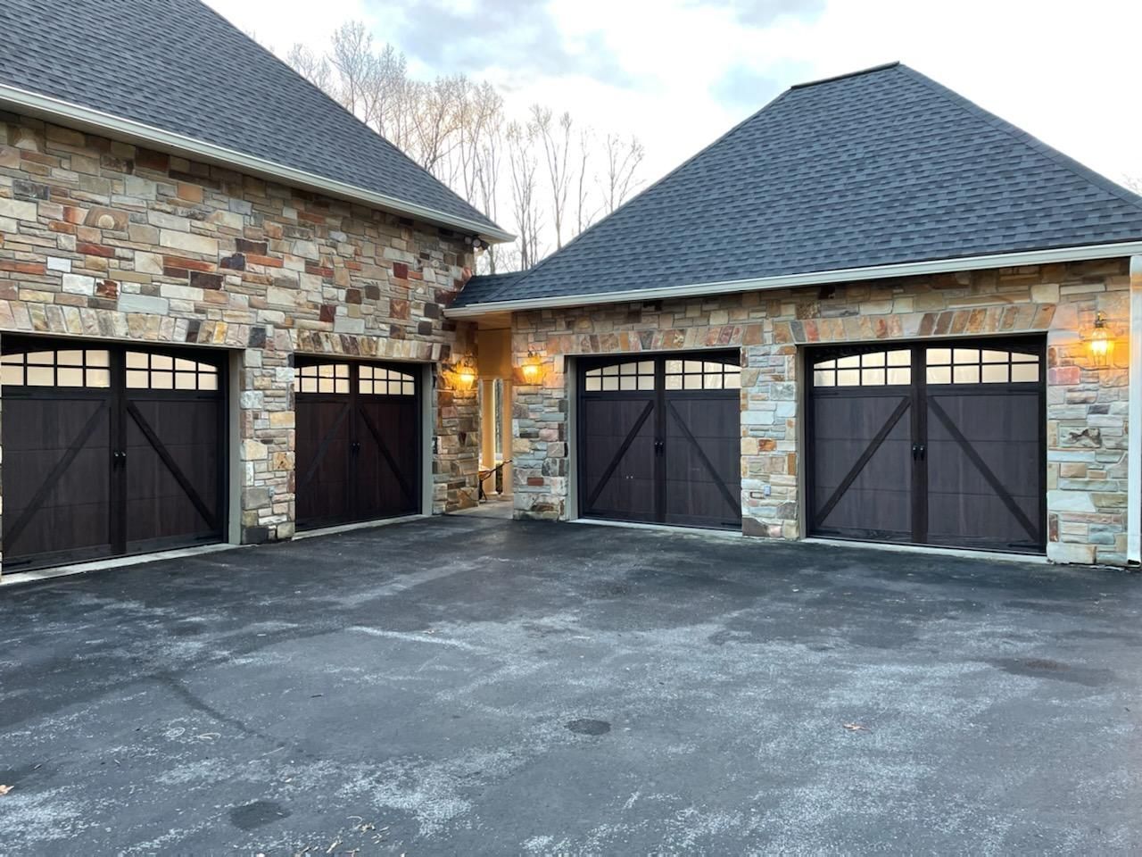 A stone house with four dark garage doors featuring decorative X-bracing and windows, set against a gray asphalt driveway.