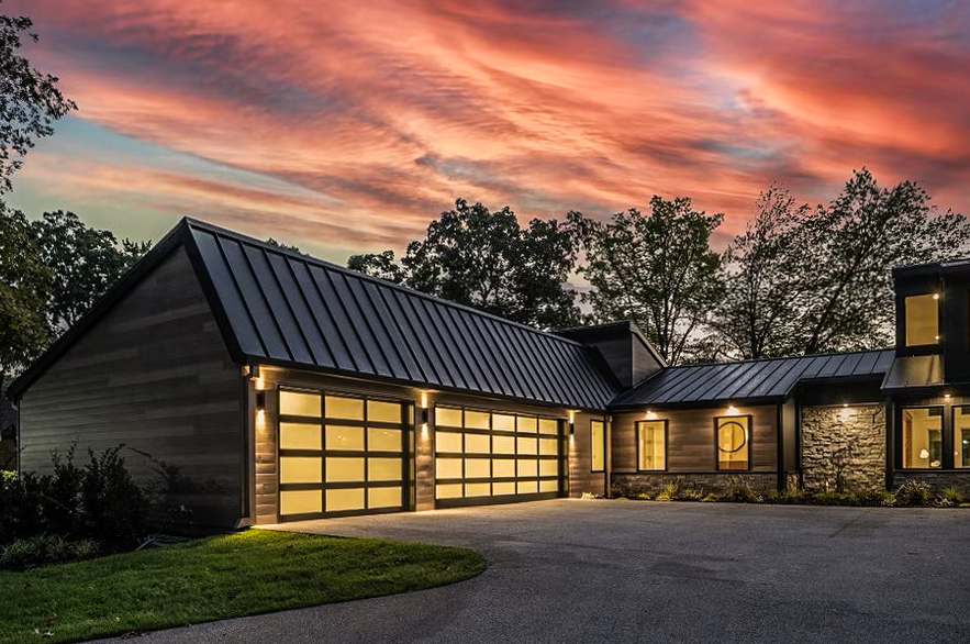 Modern house with a metal roof and large glass garage doors at twilight, set against a colorful, dramatic sky.