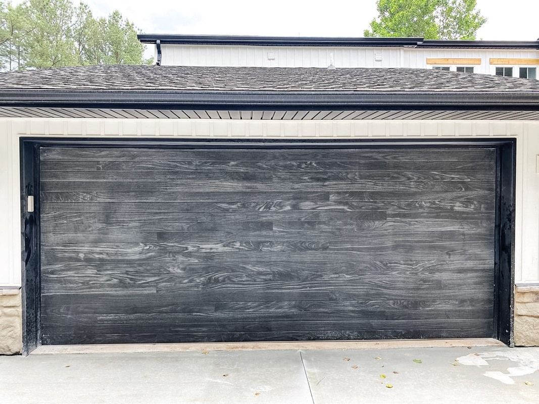 A wide, dark gray garage door with a wood-grain texture and black trim, set in a white exterior wall.