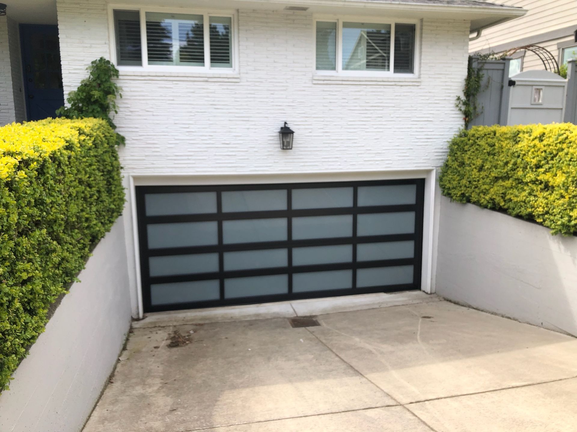 Black and frosted glass garage door with black trim in a white brick building. Green bushes frame the entrance.