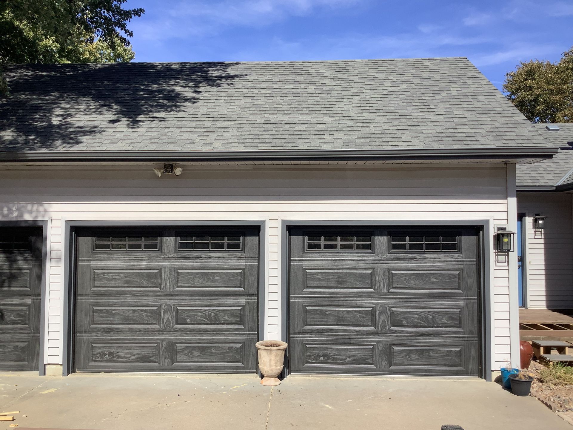 A light-sided detached garage with two dark brown garage doors featuring rectangular windows under a gray shingled roof.